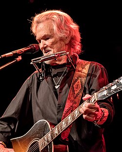 An old man with grey hair and beard, wearing a black shirt, playing a guitar