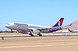 A white twin-engine plane painted with the word "HAWAIIAN" in the front and a woman in different purple hues on the tail has just landed on a runway on a sunny day amid a desert backdrop