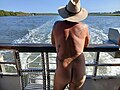 Back view of naked person in a wide brimmed hat, looking over the stern rail of a boat, with the wake going into the distance, grass covered bank on the left and trees on the right at the edge of the waterway, clear blue sky