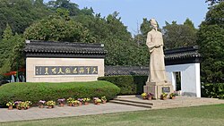 A large statue of Gu Yanwu holding a scroll in front of a sign and foliage
