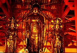 Front view of three standing statues which are covered in gold. The central statue is about two heads taller than the two falnking statues. It has the right hand pointing down with the palm towards the viewer and the palm of the left hand turned upward with the thumb touching the middle figer. All three statues have halos behind their heads with emanating rays.