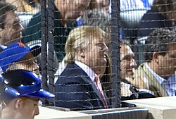 Trump, in a suit, sits in a crowded baseball stadium