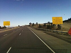 A highway near the top of a ridge. On either side of the highway are big yellow signs reading, "Trucks, Don't be Fooled—4 more miles [6.4 km] of steep grades and sharp curves".