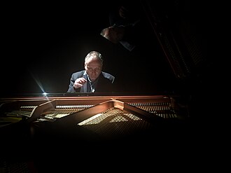 Mark Springer wearing a suit, seated behind a grand piano and appearing to play, on a darkened stage