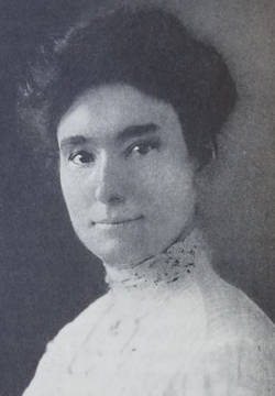 A white woman with dark hair, eyes, and brows. She is wearing a high-collared white lace blouse, and her hair is in an updo.