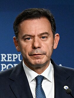 Photograph of Luís Montenegro in a dark suit, white shirt, and patterned blue tie speaking at a podium against a blue backdrop, facing forward.