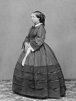 A woman in her 40s dressed in 19th century clothing, standing and looking to one side