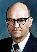 Color photo of a bald man wearing glasses and a suit with a striped tie