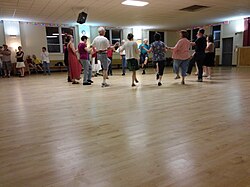 A small circle of dancers with one foot raised hold hands while several people look on.