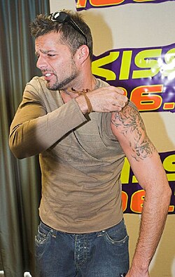Ricky Martin looking to his right, while pulling up his the left sleeve of his t-shirt to reveal a tattoo.