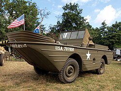 Color photograph of a vehicle on land. It looks like a boat with a prow but it has four truck wheels (only three are visible). It has a windshield and canvas covering behind it to form a cabin. It is painted in military green and decorated with white stenciled letters: "U.S.A. 707S5S" on the side of the prow, "[...] 60STD RCN46" on the rip of the prow and a big white five-point star on the side. It has a rear-view mirror out of the side of the "cabin".