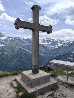 Big Summit Cross made of Stone on a Mountain Peak with some alpine peaks in the background