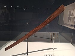 What appears to be a somewhat flat wooden stick with a slight curve, held up by a vertical metal bar in a glass case. Additionally, four Obsidian blades are also displayed next to the Macuahuitl.