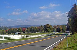 A rolling landscape with hills in the distance, a line of trees some of which are turning red, and fields in the foreground. A white fence separates them from a road that stats at bottom left and then passes between trees at the right center before cresting. At the bottom right is a small green sign with white numbers on a metal stand.