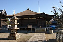 Wooden building with a pyramid shaped roof and an open veranda on the front. A stair leads at one end of the veranda to the building's level.
