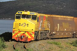 Yellow diesel locomotive hauling brown hopper wagons with the ocean and a large hill in the background