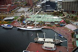 A view of a brick promenade with a green-roofed shopping mall and the USS Constellation. Two other boats are in front of the Light Street Pavilion, and it also features a green sea monster wrapped around one of its balconies (Ripley's Believe It or Not!) On the left of it are blue awnings for the Bubba Gump Shrimp Co. restaurant, and on the right, that signage of a blue M is for McCormick World of Flavors. On the far right of the promenade is an ice rink and the amphitheater.