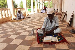 A dark-skinned boy sits cross-legged on a small mat on a tiled floor in a shaded colonnade. He is wearing light clothes and a white cap and is looking down at a book written in Arabic. Beside him is a tin can. Some distance behind him are more boys sitting and standing.