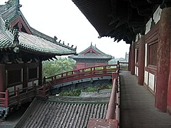 Several wooden buildings with grey tile roofs, connected by an arched bridge. A forest can be seen behind the buildings.