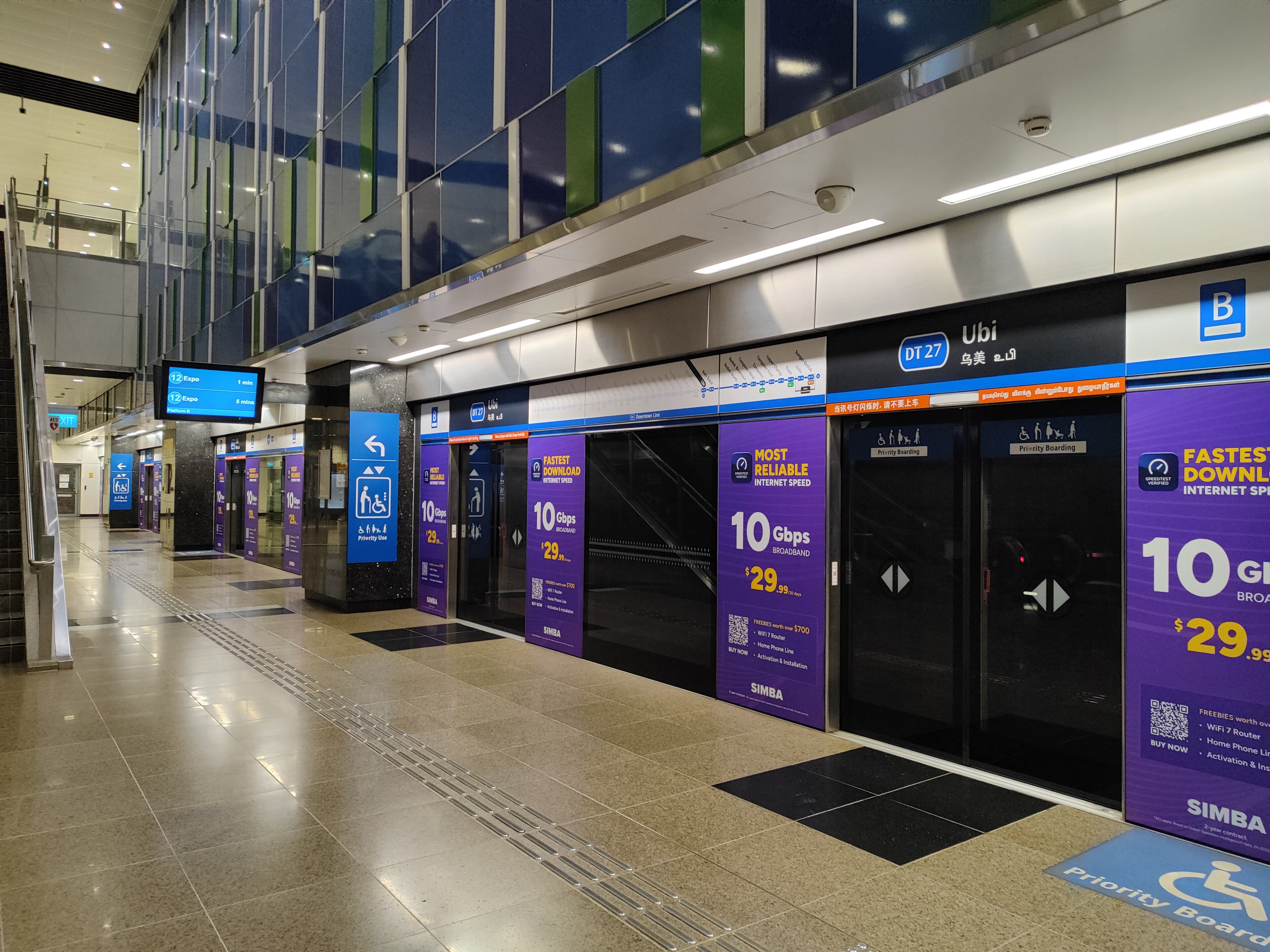 Ubi underground platform featuring blue and green vertical accent panels on the far wall. The station has a spacious feel with recessed circular ceiling lights.