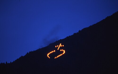 A photograph of a dusk scene. On the top, left, a dark blue triangle of dusk sky. On the bottom left, a black triangle of distant hillside. On the hillside, several fire points form a schematic round shape crowned by a Latin cross.