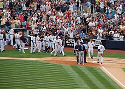 A man in a white baseball uniform with navy pinstripes and the number "2" on the back of his uniform runs towards home plate, while his teammates run to meet him to celebrate.