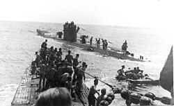 Three-quarter front view over the bow from a submarine conning tower of another submarine with numerous people standing on both submarines, at sea.
