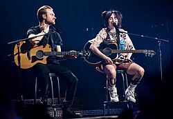 Billie Eilish and Finneas O'Connell seated at center stage, holding their acoustic guitars
