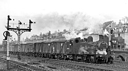 A black-and-white busy photo of a goods train in a yard, producing a lot of smoke. There are houses behind elevated above the railway. There are semaphore signals on the adjacent tracks and one track in the background is elevated on a brick viaduct