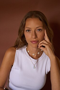 Portrait of a young woman with long hair and a direct stare. She is resting her face in her left hand in a thoughtful manner.
