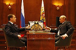 Medvedev and Putin, both white men, wearing suits sitting at a wodden desk across from one another. There are flags in the background.