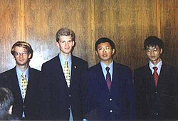 Four boys in black suits with bluish-white dress shirts and brightly-coloured ties standing in front of a wall composed of wooden panels