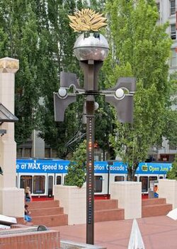 A color photograph of an urban center area.  A tall, thin structure stands adorned with various lights and appendages, with a globe-like object on top.