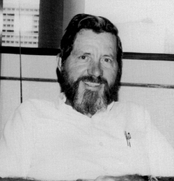 A smiling middle-aged man with a greying beard in a white button down shirt with pens in the pocket