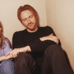 A sullen bearded ginger man holds someone's hand off-frame as he sits in the corner of a room.